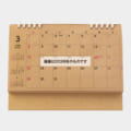 Wooden desk calendar open to March 2026, with a small left-side mini calendar and a white banner containing Japanese text across the center.