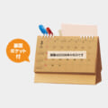 Wooden desk calendar with a back pocket; orange speech bubble reads '裏面ポケット付' (back pocket included). Calendar shows 2026 and a note in Japanese about the year.